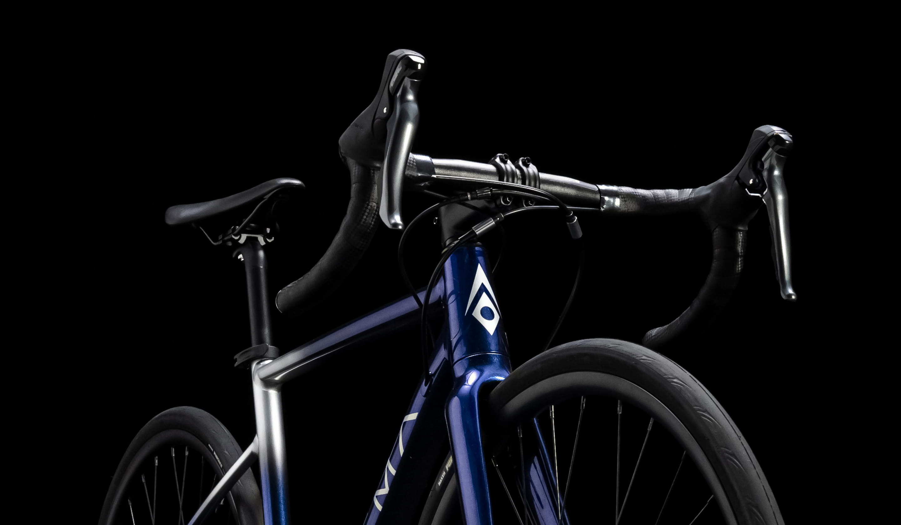 黒い背景に、ロードバイクのハンドルとフロント部分が強調された構図の写真。青とシルバーのフレームに、カーボンハンドルバーやディスクブレーキが特徴的で、スポーティかつ洗練されたデザインが際立つ。